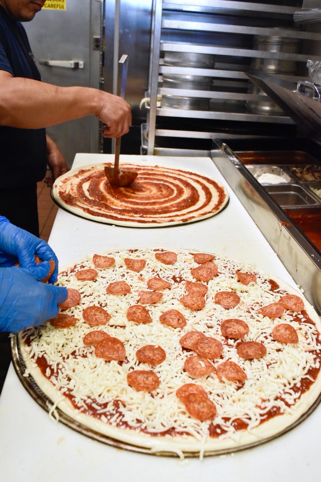 Photo of a man spreading pizza sause onto a pizza an another man placing toppings on a pizza